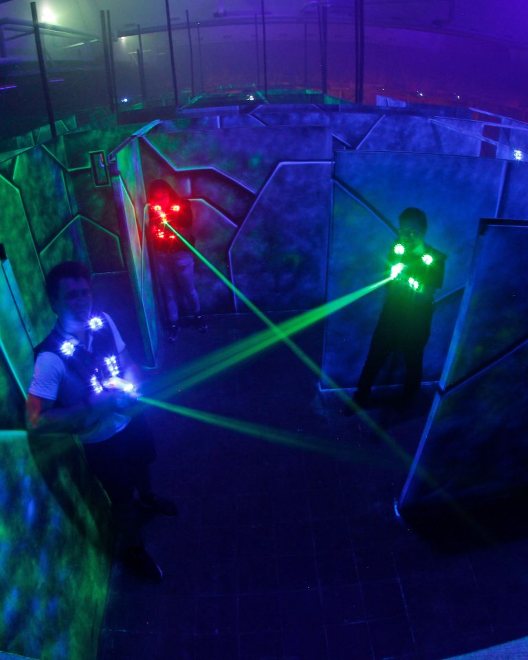 Drei Personen in unterschiedlich leuchtenden Westen stehen in einer großen futuristisch gestalteten Lasertagarena