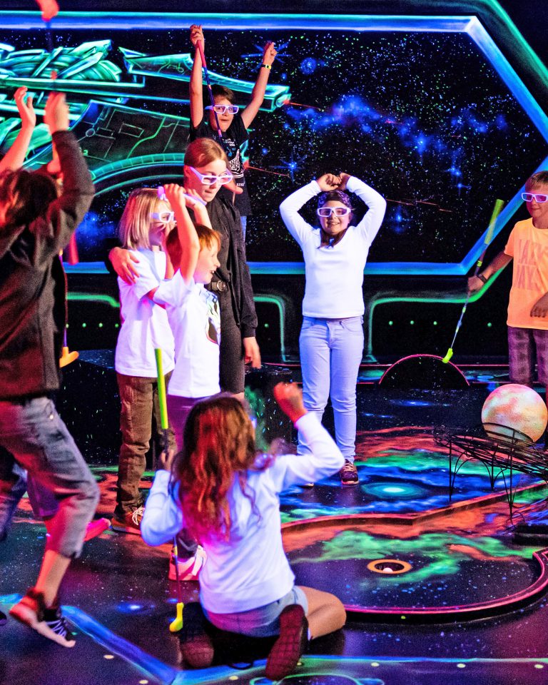 Eine Gruppe Kinder jubelt einen Jungen an, der grade den Ball auf einer Minigolfbahn im Weltall-Raum eingelocht hat.