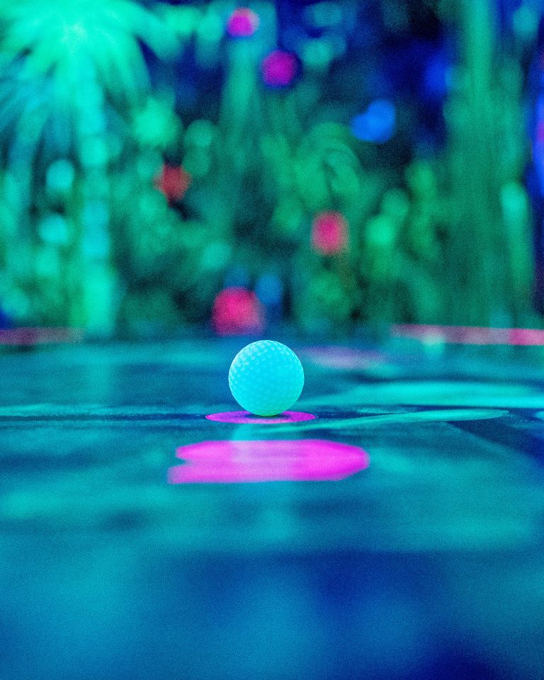 Nahaufnahme eines Minigolfballs auf dem Startpunkt einer verschwommenen Minigolfbahn im grünen Dschungelraum von NeonGolf.